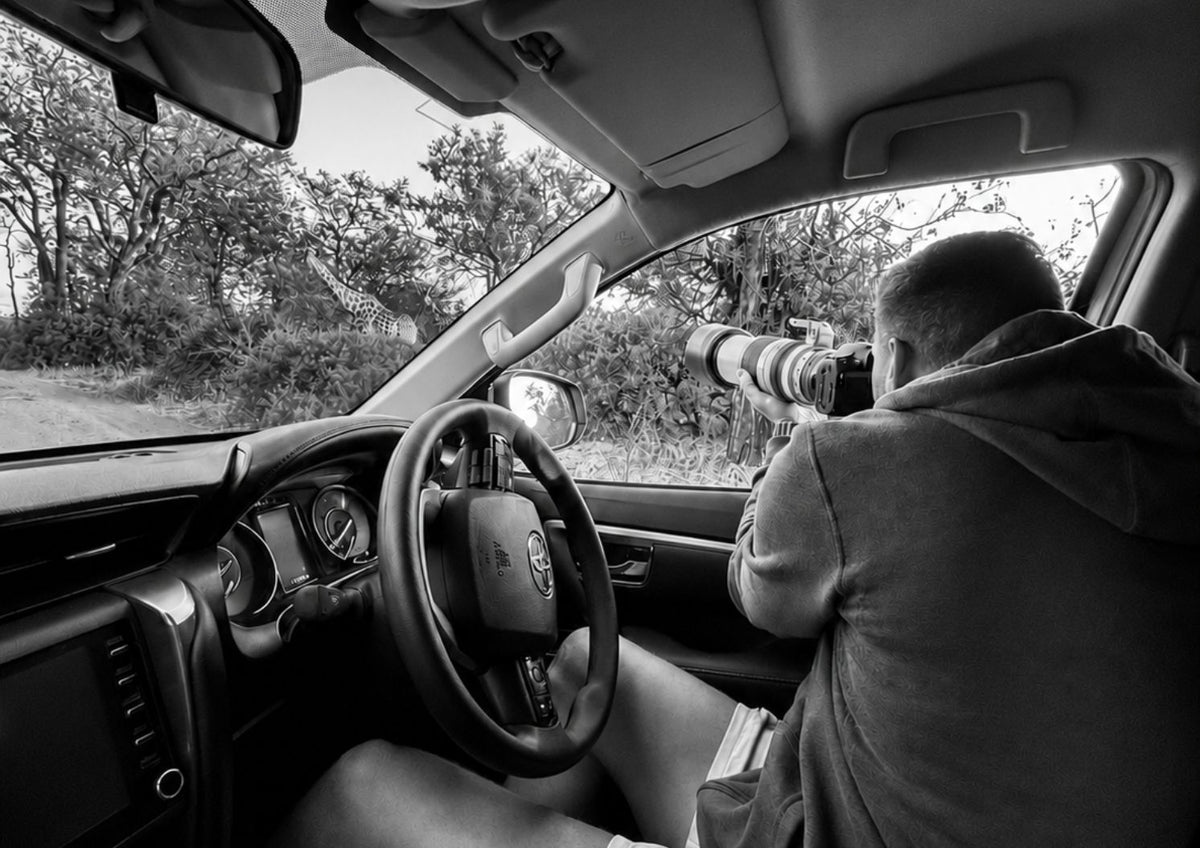A black and white photograph inside a car, showing Dan Koopowitz holding a large telephoto camera lens to photograph a giraffe in the Kruger National Park.
