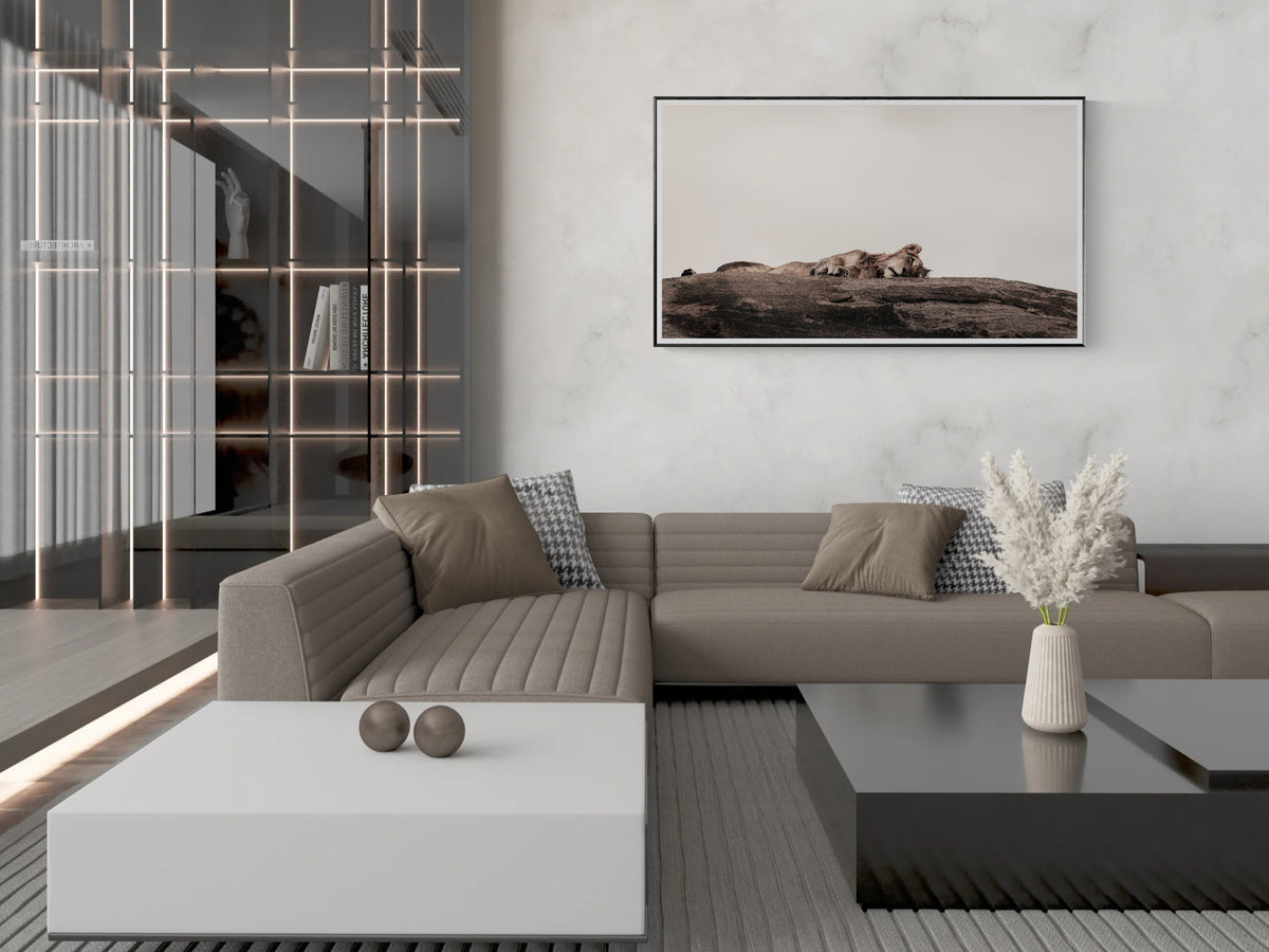 A modern living room featuring a large framed art print of Dormant Ridge on a light gray marble wall above a taupe sectional sofa. The room includes a white coffee table, a black side table with a vase of dried grass, and a glass shelving unit with warm metallic accents.