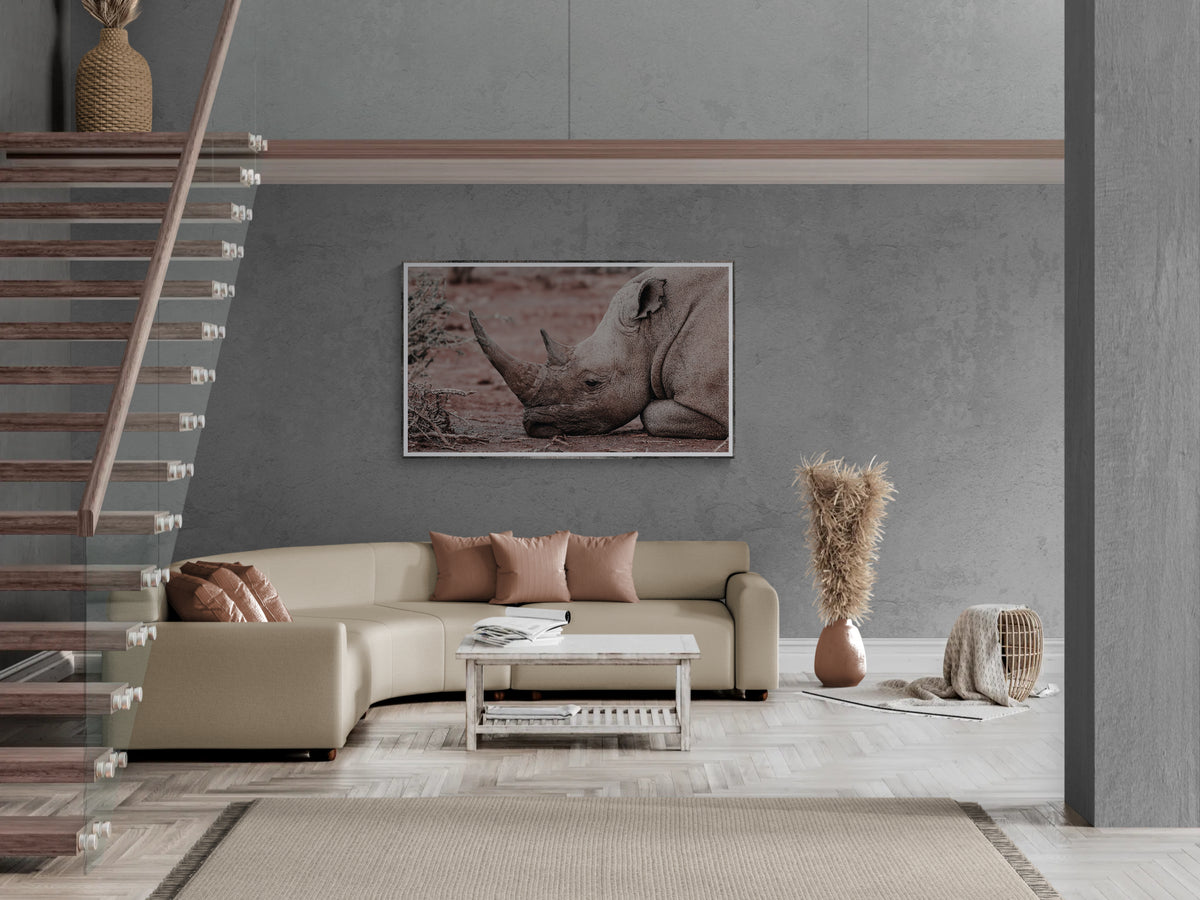 A framed horizontal art print depicting a rhinoceros in muted earthy tones, displayed on a textured gray wall in a modern living room setting with a beige sectional sofa and staircase.
