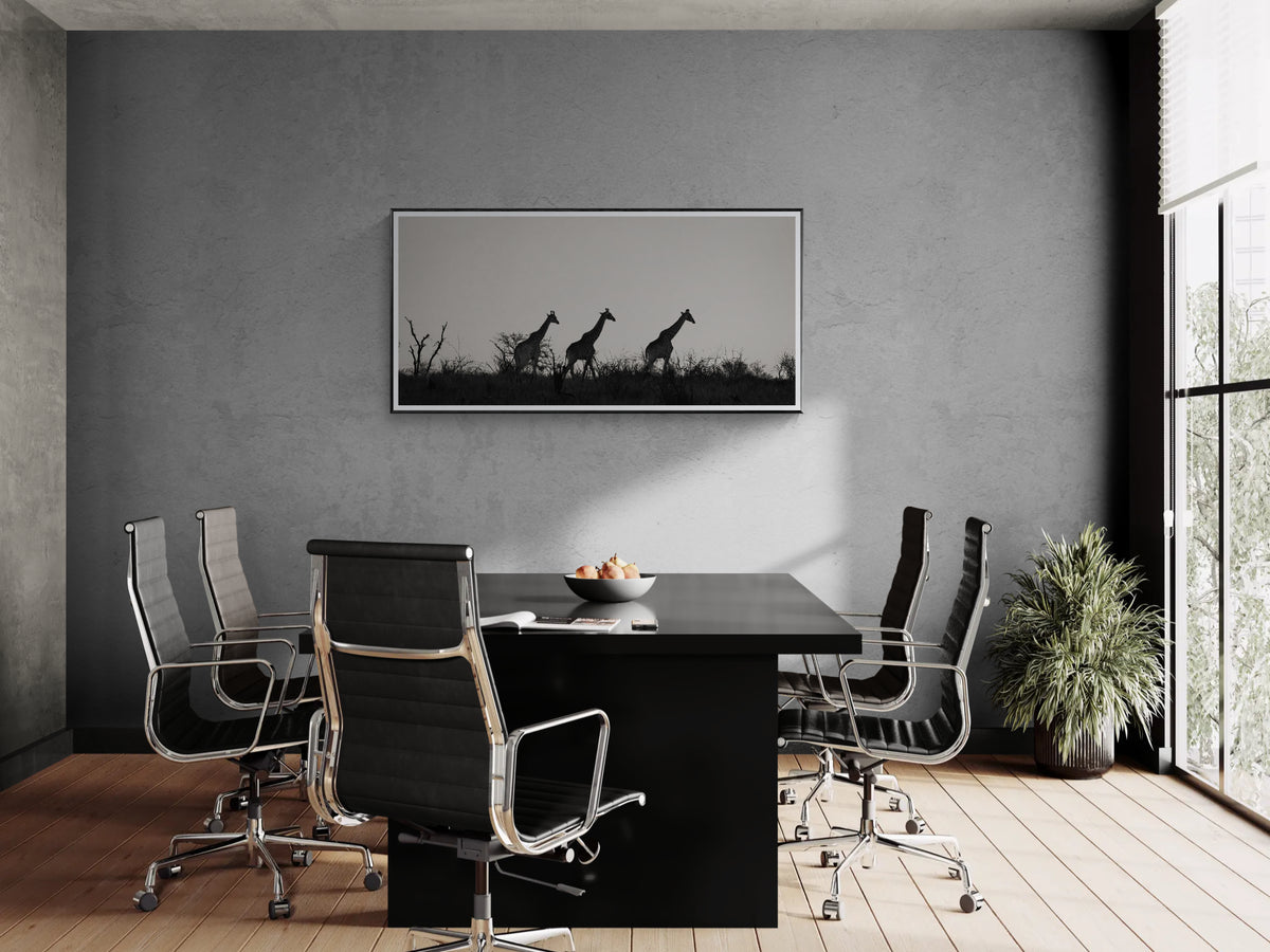 A modern conference room with a black rectangular table and six black mesh office chairs with chrome bases. On the textured grey wall above the table is a framed black-and-white wildlife fine art print depicting silhouetted giraffes.