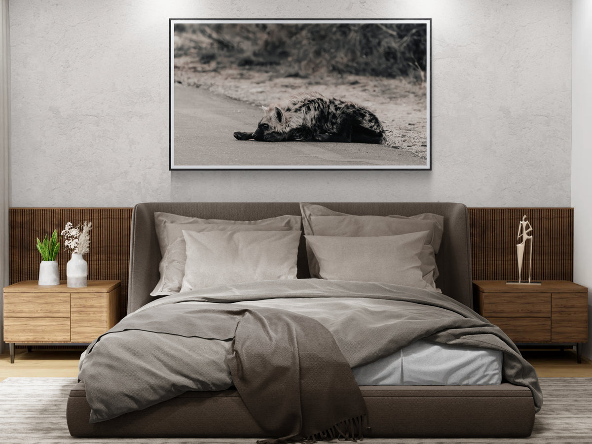 A modern bedroom featuring a large black and white framed art print of Grounded depicting a hyena on a road, hung above a low-profile platform bed with neutral grey bedding and wooden nightstands.