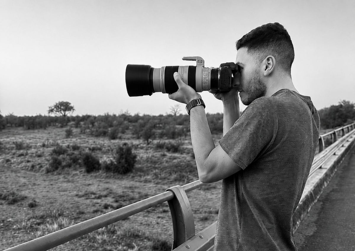 Dan Koopowitz holding a large professional camera with a long telephoto lens, looking through the viewfinder outdoors on a bridge overlooking a dry riverbed in Kruger National Park.
