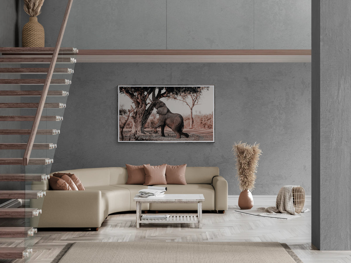 A framed art print depicting an elephant under a tree, displayed on a gray wall in a modern living room setting with a beige sectional sofa and staircase.