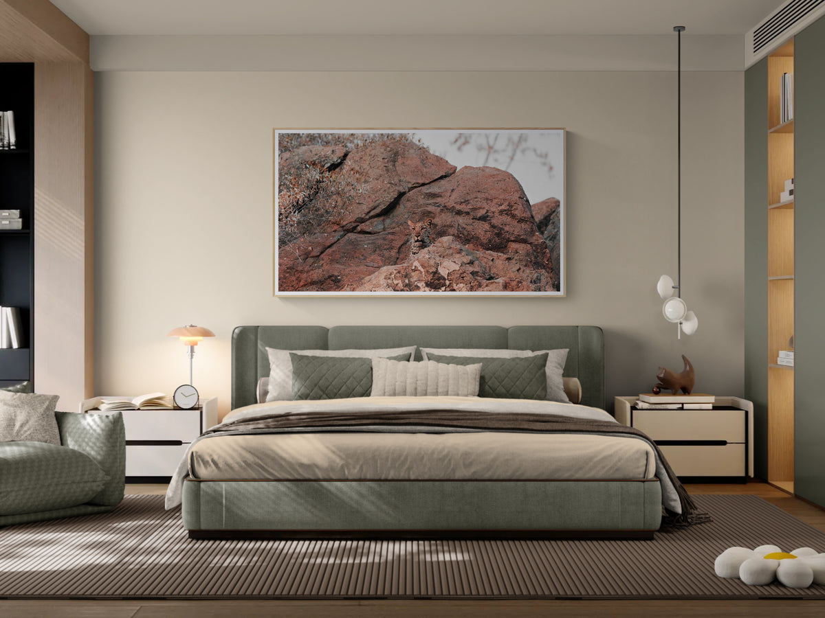 A modern bedroom featuring a large framed landscape fine art print of reddish-brown rock formations on the wall above a green upholstered bed. The room is styled with neutral beige tones.