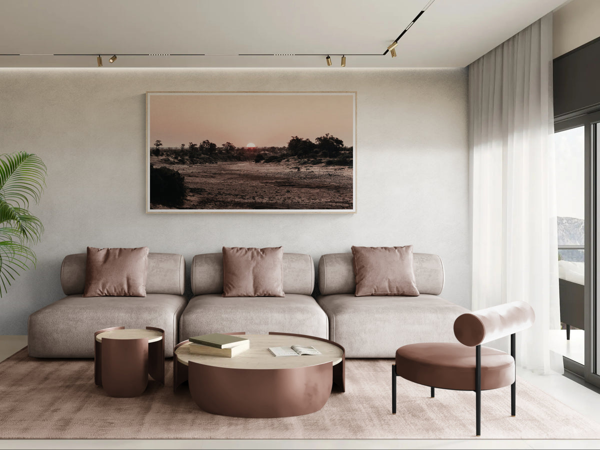 A modern living room featuring a large framed landscape fine art print on a textured light wall above a beige sectional sofa. The print depicts a dry field landscape at sunset with warm brown, beige, and black tones.