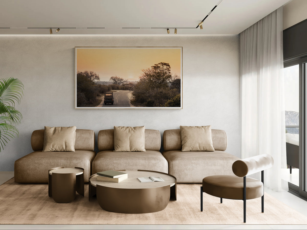 A framed art print of Trailing The Sun depicting a sunset over a rural road is displayed on a textured light grey wall above a beige sectional sofa in a modern, minimalist living room.