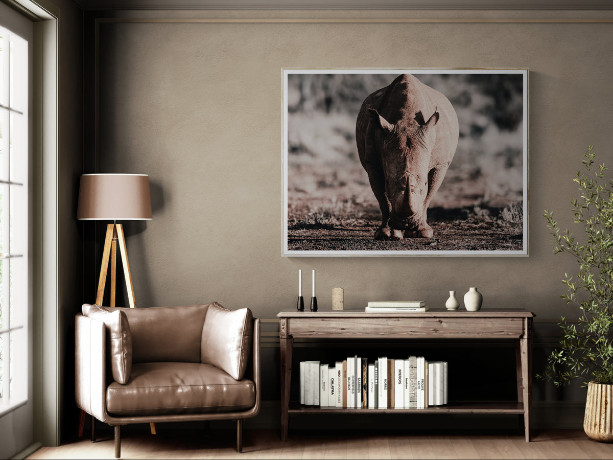 A framed wildlife photograph of a rhinoceros hanging on a taupe wall in a styled living room setting with a leather armchair, console table, and floor lamp.
