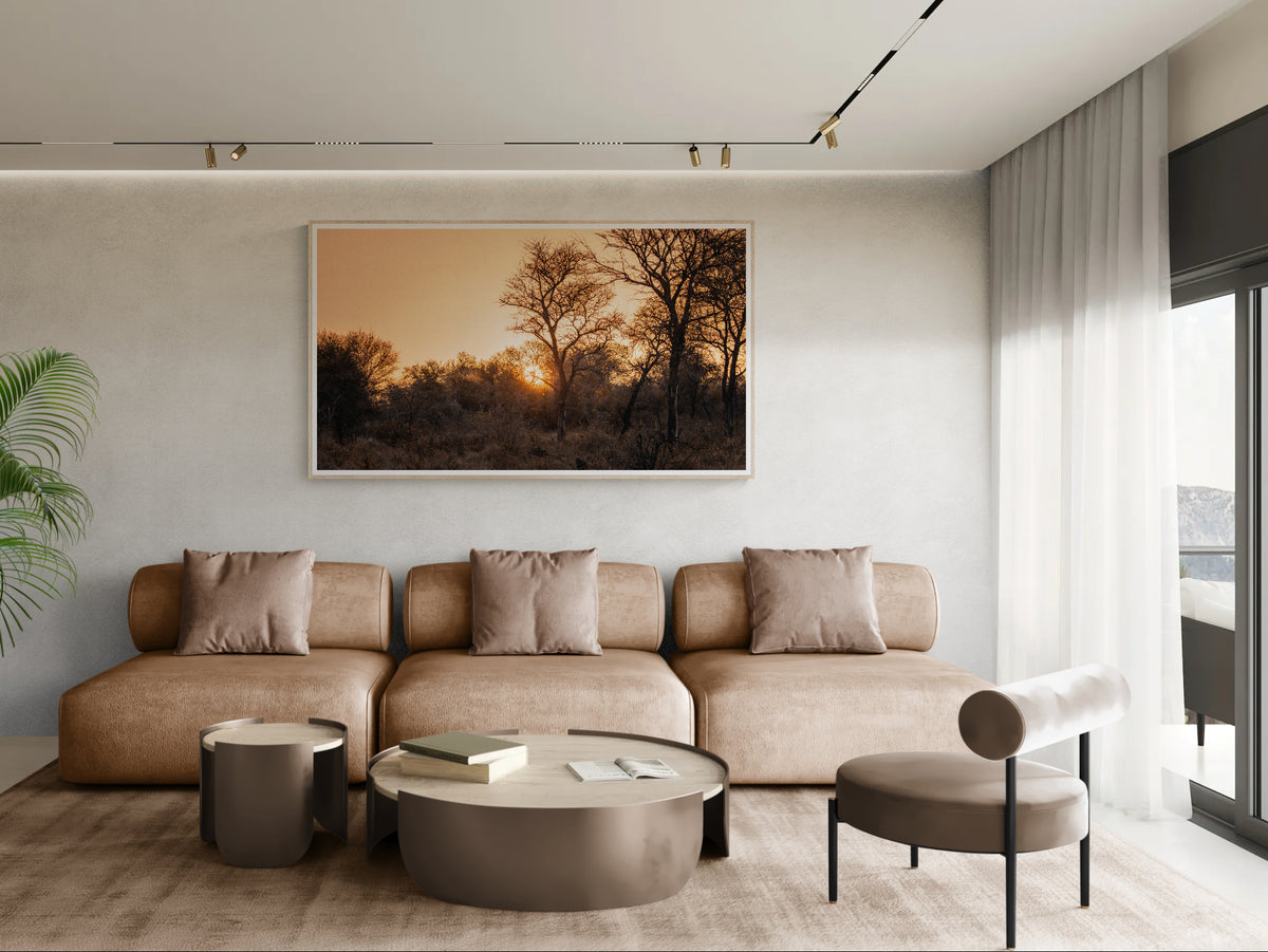 A large framed art print of Veld Gold depicting a sunset over trees, displayed on a light gray wall above a beige sectional sofa in a modern, minimalist living room.